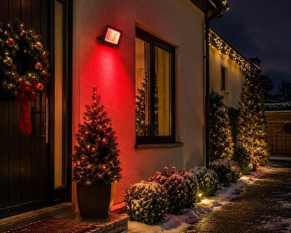 A house front at night washed in red light from an RGB floodlight, with a Christmas tree by the door